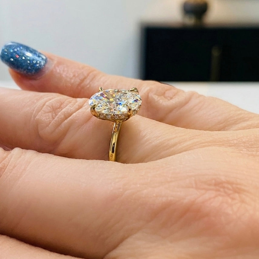 A 2 carat oval-cut lab-grown diamond engagement ring set in a slim 14K yellow gold band. The side profile reveals a delicate hidden halo of small diamonds encircling the base of the center stone