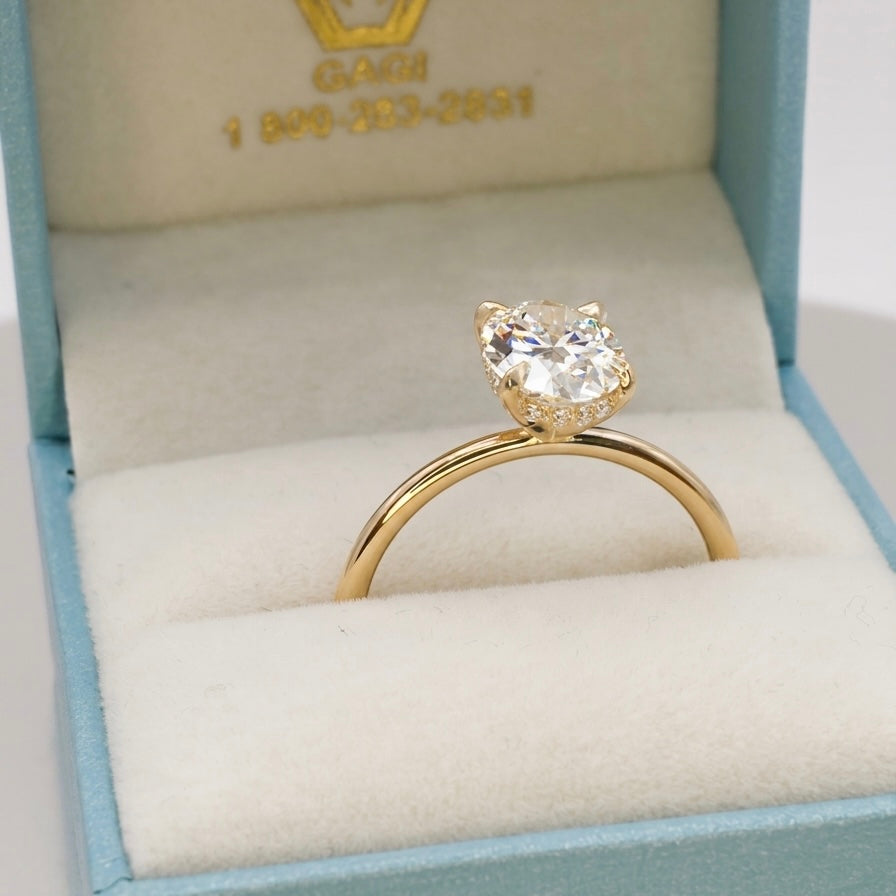 A 2-carat oval-cut lab-grown diamond engagement ring set in a slim 14K yellow gold band in a gift box. The side profile reveals a delicate, hidden halo of small diamonds encircling the base of the center stone